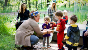 An adult wearing a crown of paper an autumn leaves crouches to talk to young children in a natural setting.