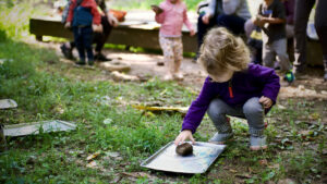 A child crouches down to work with a paper while other children play in the background outdoors.