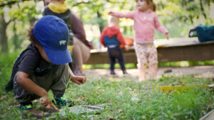 A child in a blue ballcap crouches in the grass while other children play in the distance.