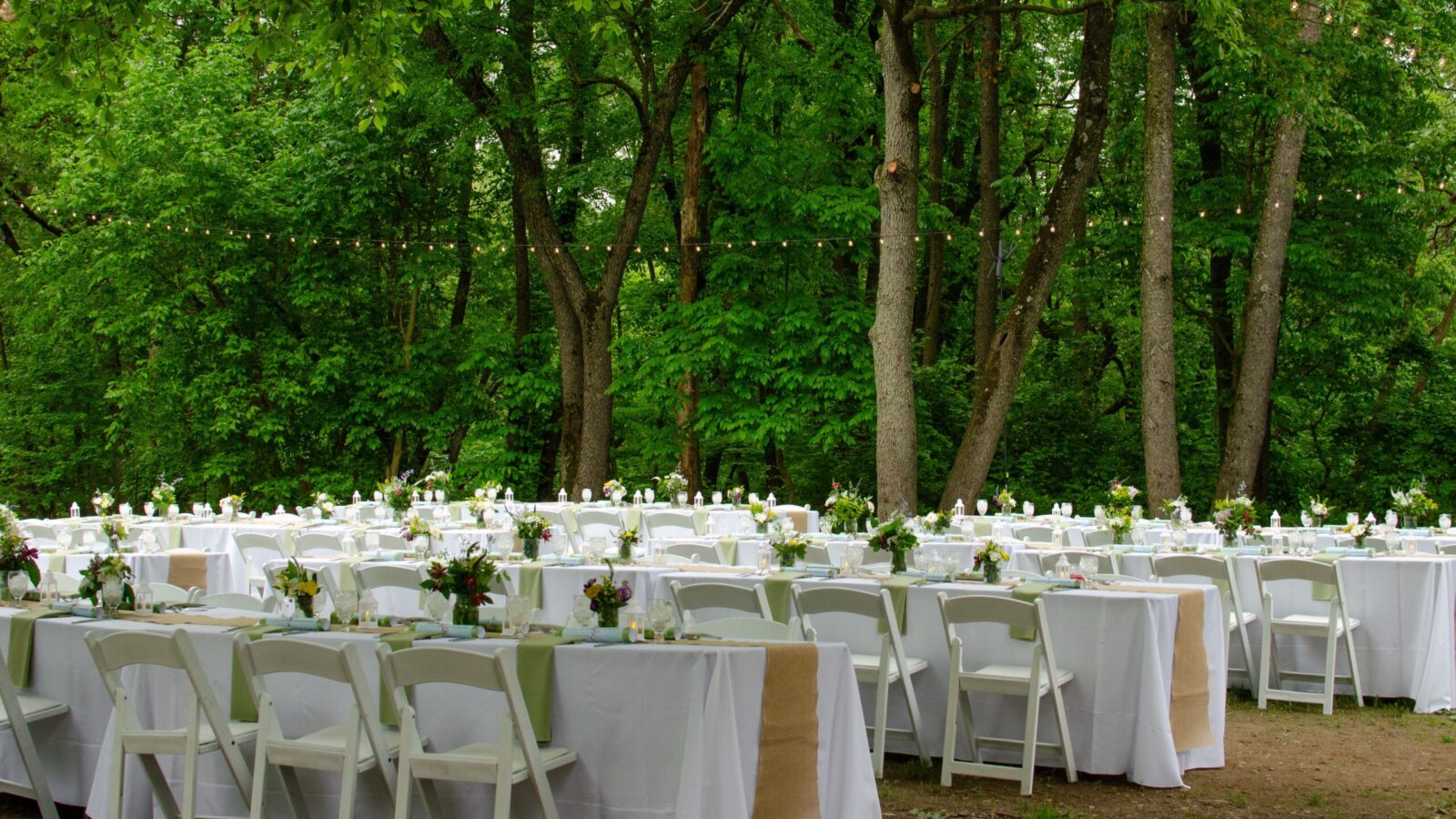 Long tables covered with white tablecloths surrounded by white chairs in a large clearing surrounded by tall trees.