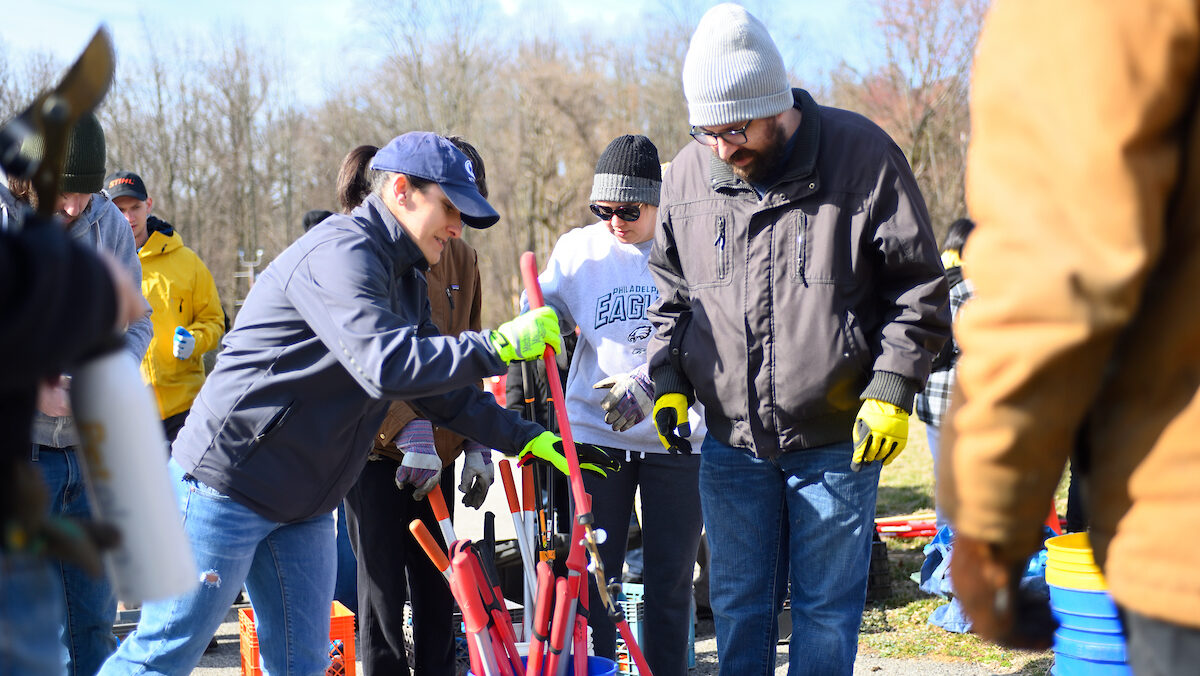 Volunteers in winter clothing gather hand tools from a bucket outdoors.