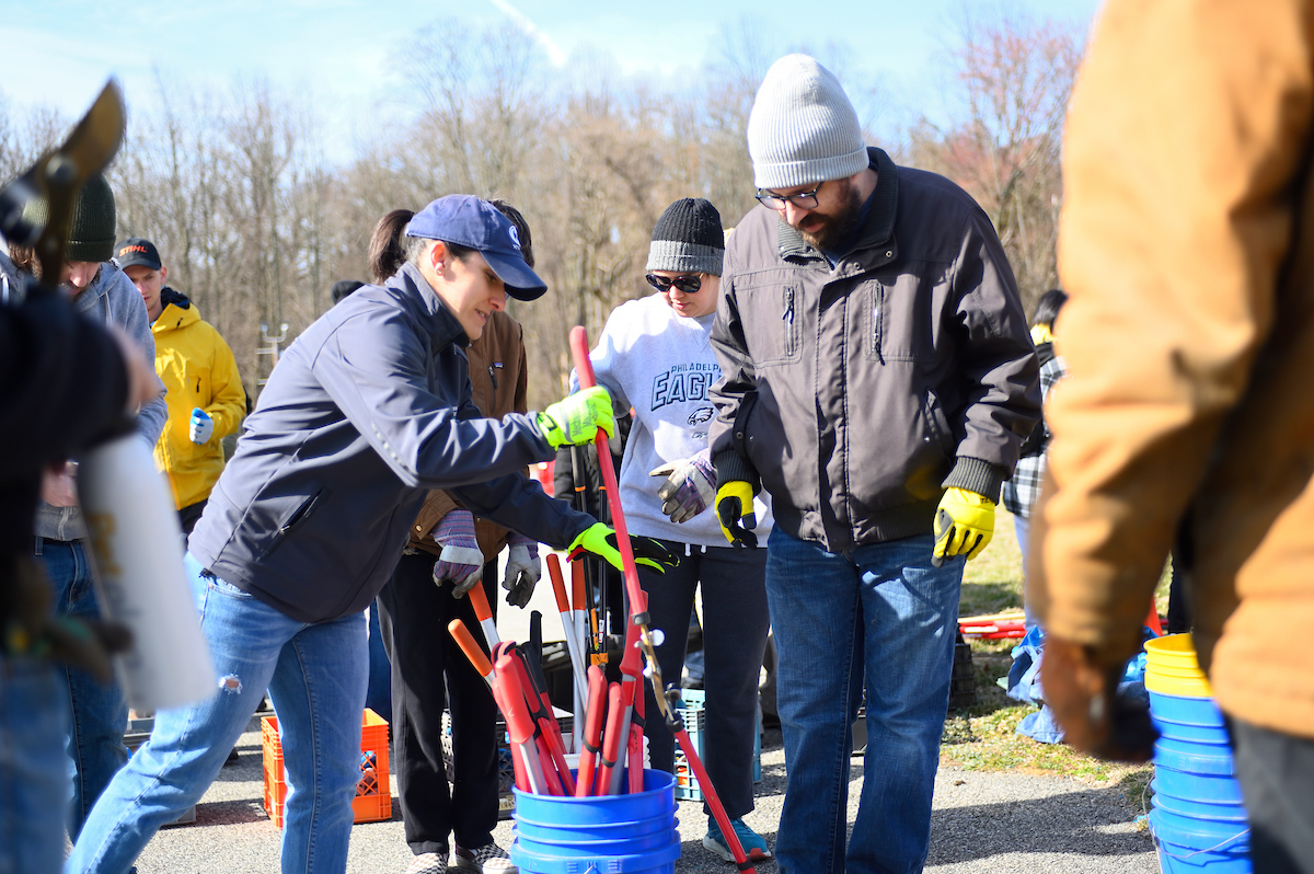 Volunteers in winter clothing gather hand tools from a bucket outdoors.