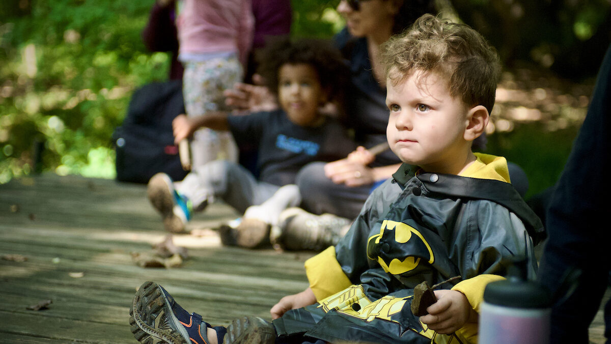 A toddler dressed in a batman costume sits on a wood platform while children and parents sit and stand in the backround.