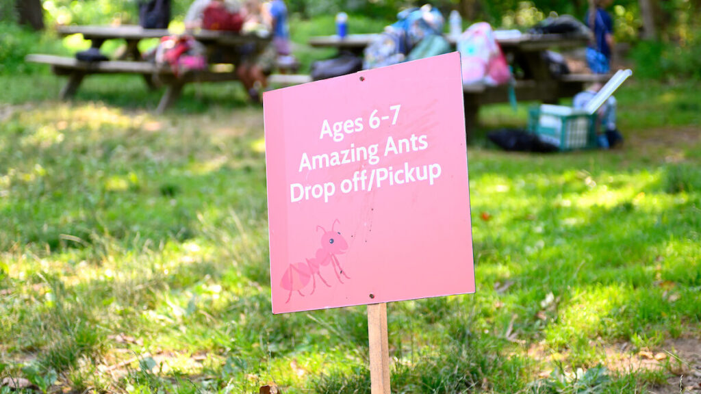 A red sign outdoors that says Ages 6-7 Amazing Ants Drop off/Pickup