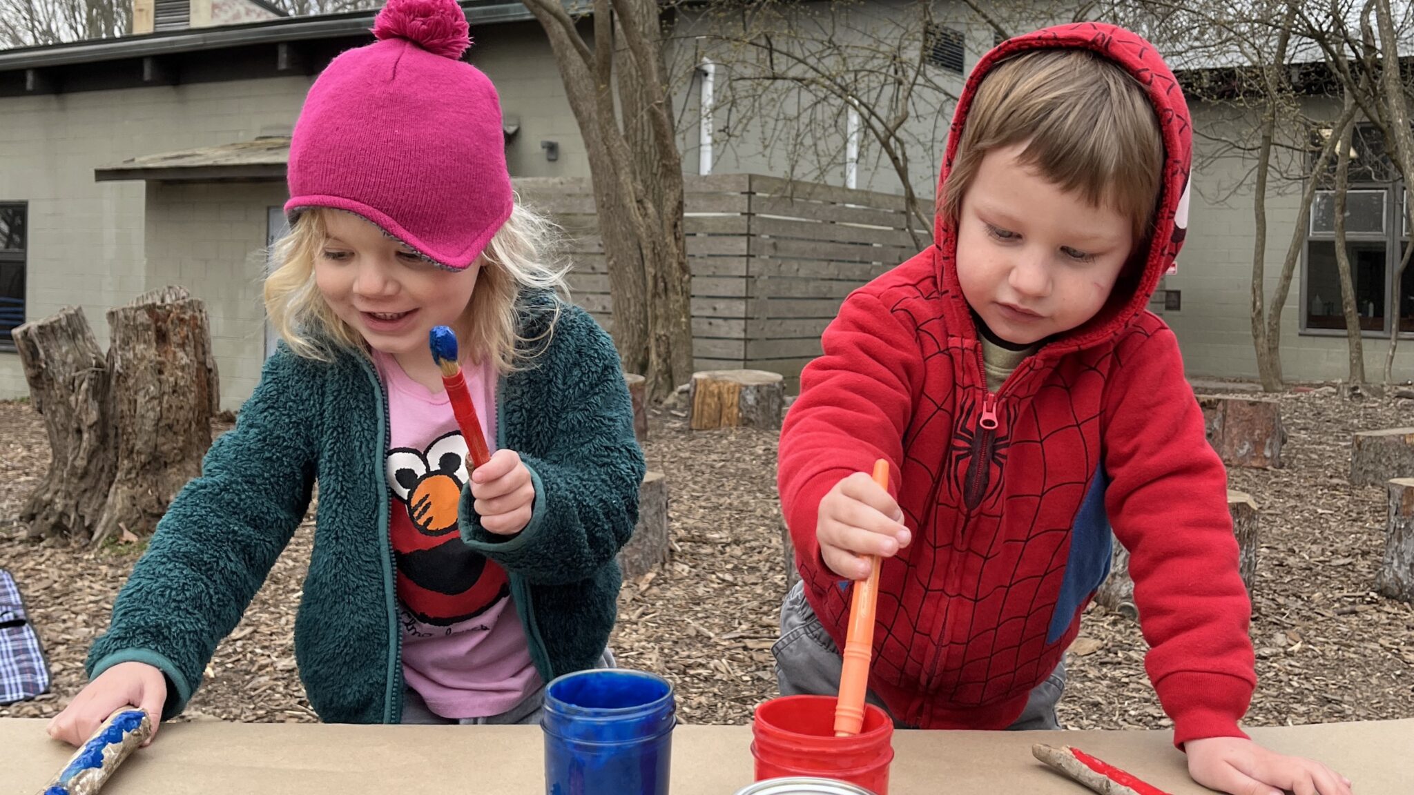 Two children in winter clothes mix paint outdoors.