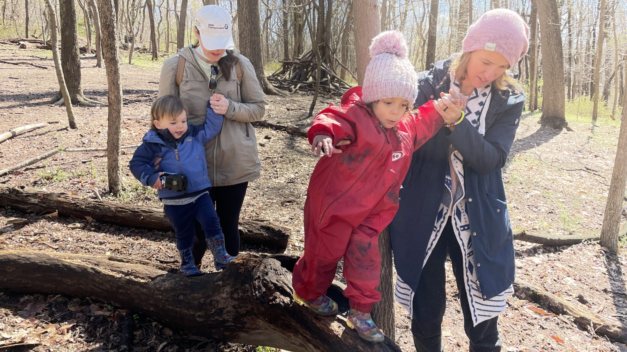 Parents hold hands with children dressed in winter clothes walking on a log outdoors.