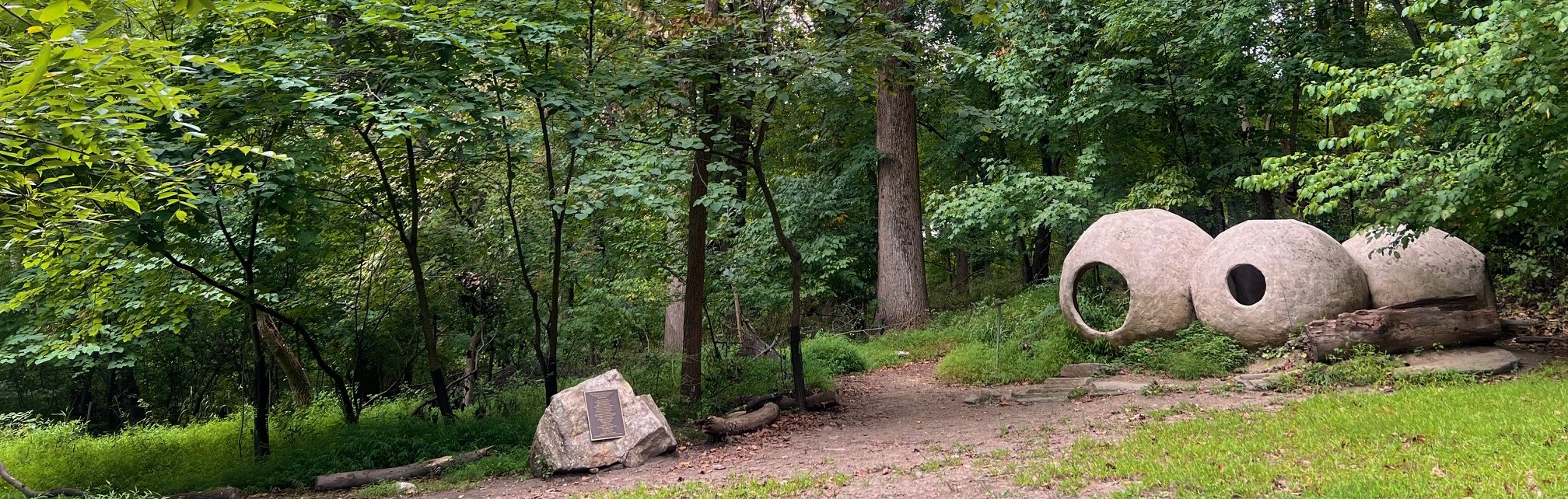 Jubilee grove at the Schuylkill Center, with two diverging trails, a sculpture of three circles and a rock with a plaque in a clearing in the forest.