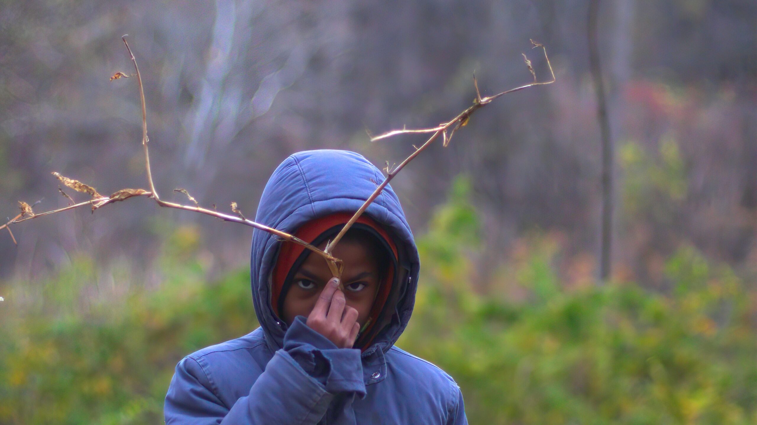 A child wearing a blue coat holds a stick in front of their face in a natural setting.