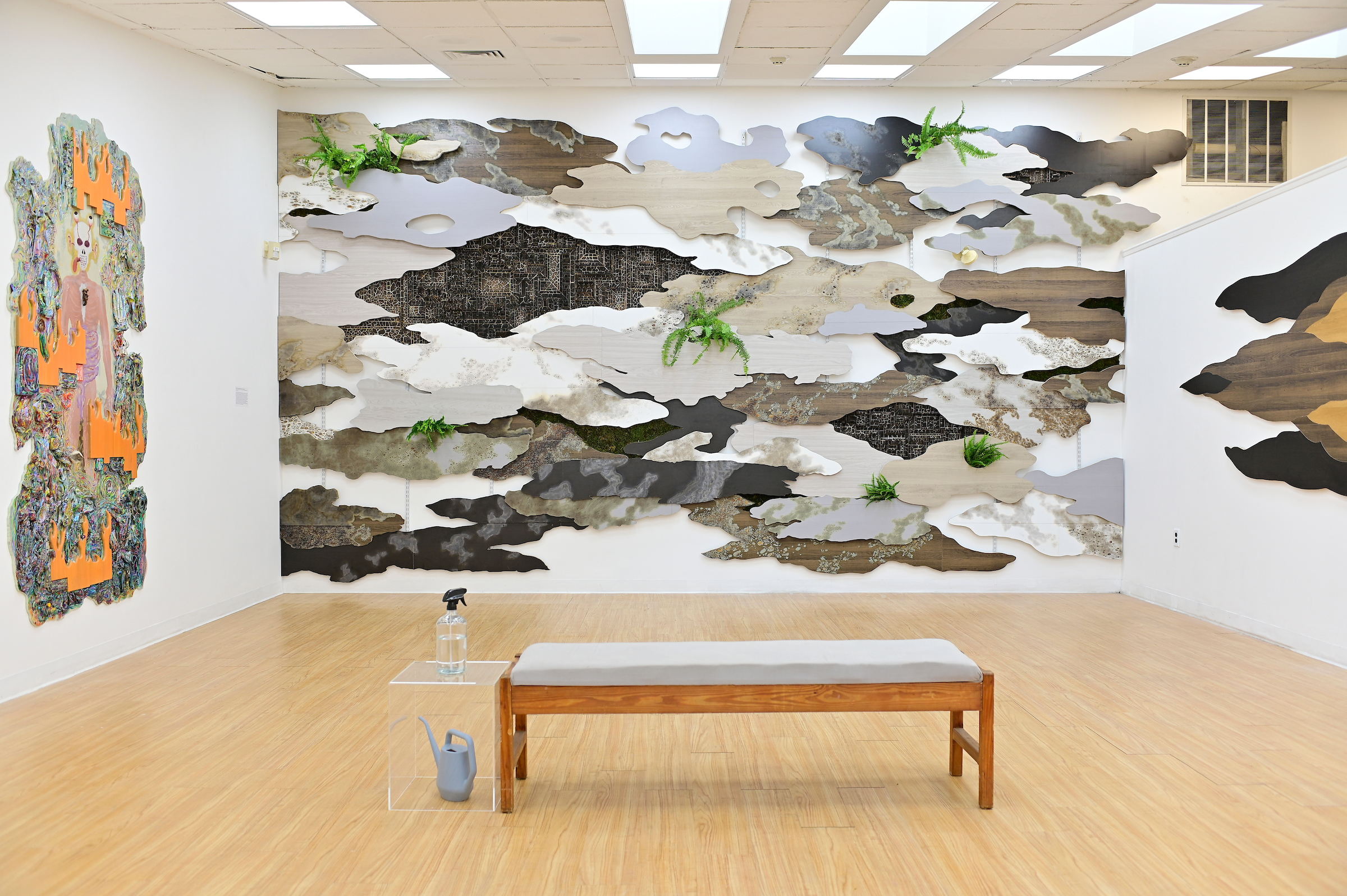 A gallery space with a wooden bench and a large wall with the shapes of grey and place clouds mounted on the wall, interspersed with small green ferns.