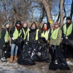 A group of volunteers wearing yellow vests hold trash bags and smile outdoors in the winter.