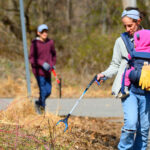 A mother holding a baby picks up trash using a trash picker while on the side of the road on a winter day.