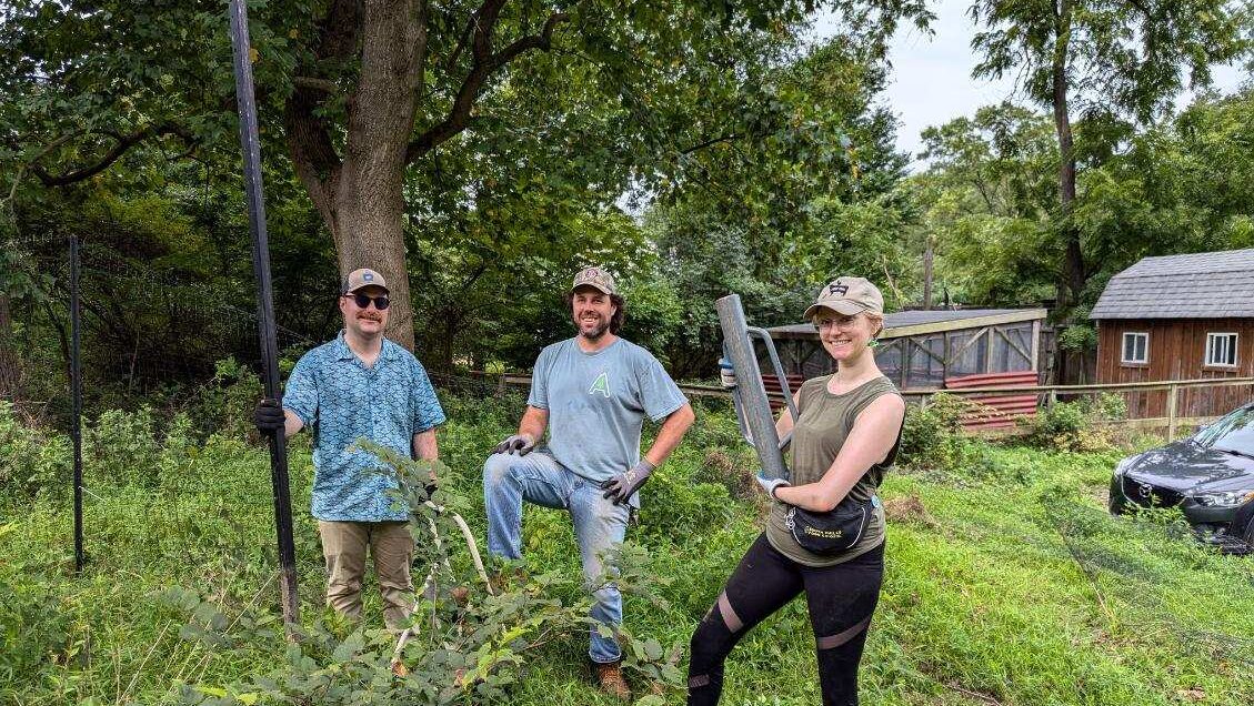 A group of three people holding hand tools stand outside smiling.