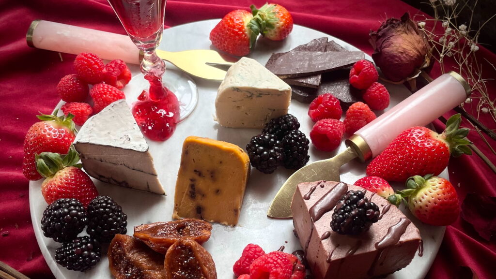 A plate of vegan cheese with fruit on a table with a red tablecloth and serving cutlery with pink handles.