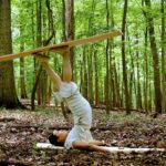 A dancer in white balances wood on their feet upside down in the forest.