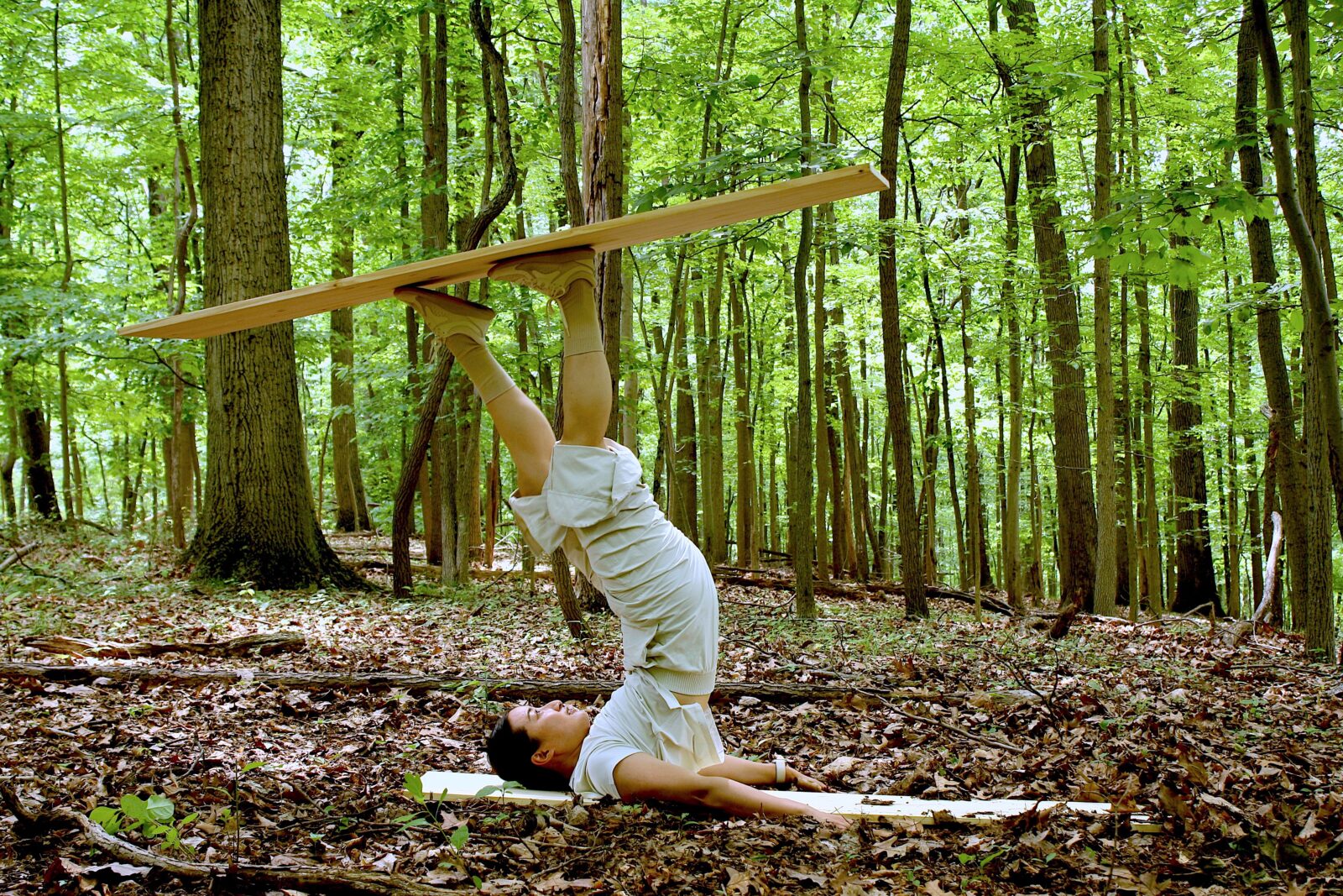 A dancer in white balances wood on their feet upside down in the forest.