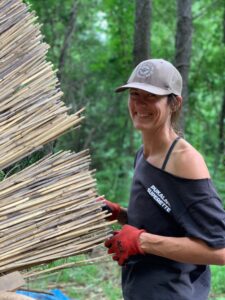 Artist Sarah Kavage works with wooden stems outdoors wearing a t-shirt, gloves, and a baseball cap. 