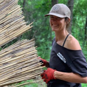 Artist Sarah Kavage works with wooden stems outdoors wearing a t-shirt, gloves, and a baseball cap.